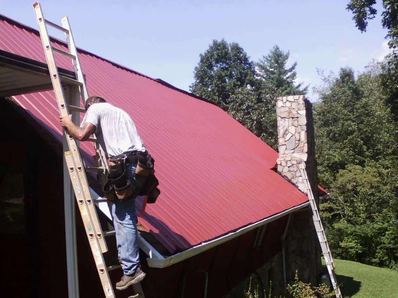 Roofer on a ladder beside a red metal roof and stone chimney — Soffit & Fascia Repair by Roofing Brothers in Hornbrook